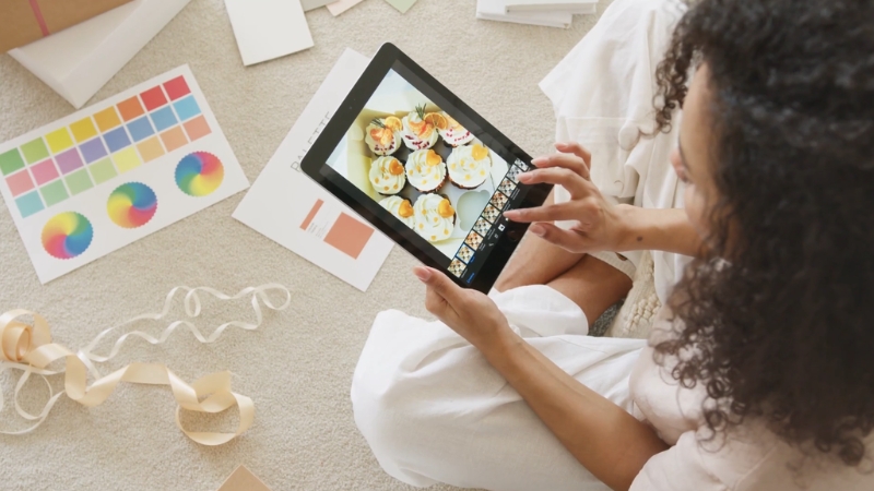 A woman edits a photo of cupcakes on a tablet while working on creative brand materials at home
