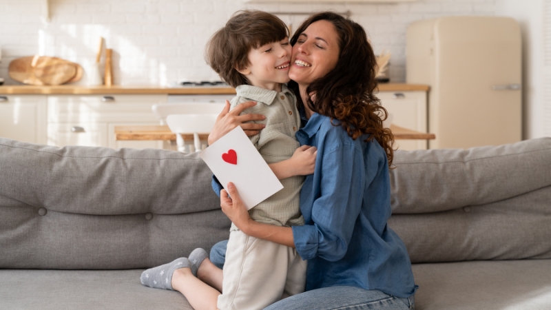 A mother and child sharing a joyful hug, with the child holding a card with a red heart for Mother’s Day