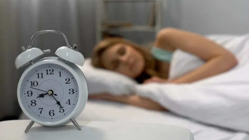 A woman sleeps peacefully in bed as a white alarm clock shows 7:00, symbolizing the impact of a consistent morning routine