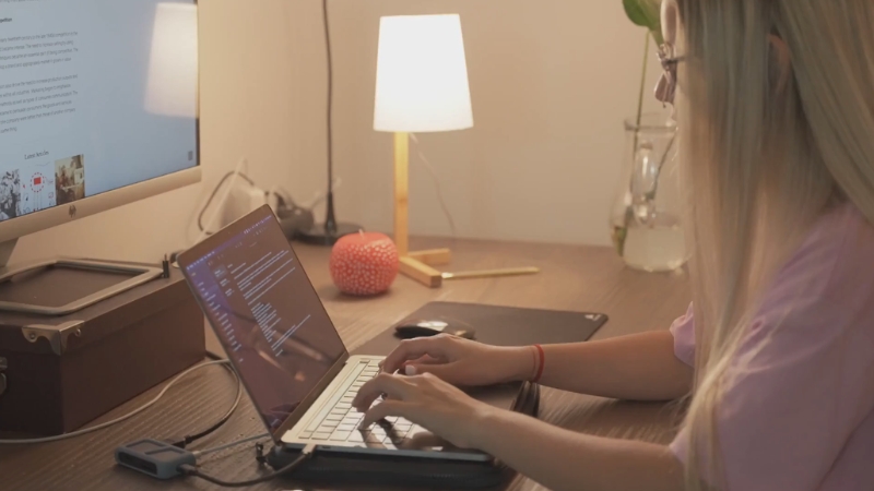 A woman types code on a laptop at a tidy desk with a monitor, lamp, and small decorations