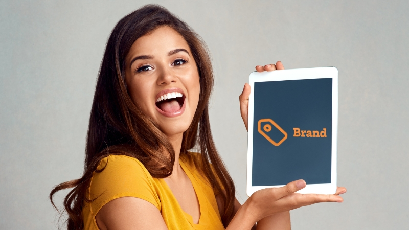 Smiling woman holding a tablet with the word “Brand,” showing how to promote a brand on a budget