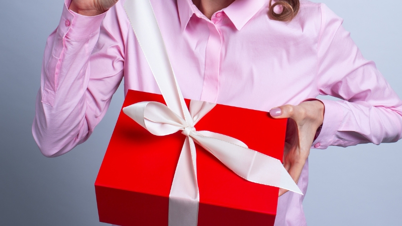 A person in a pink shirt holding a red gift box with a white ribbon, teasing it open—perfect for a post about funny gift ideas