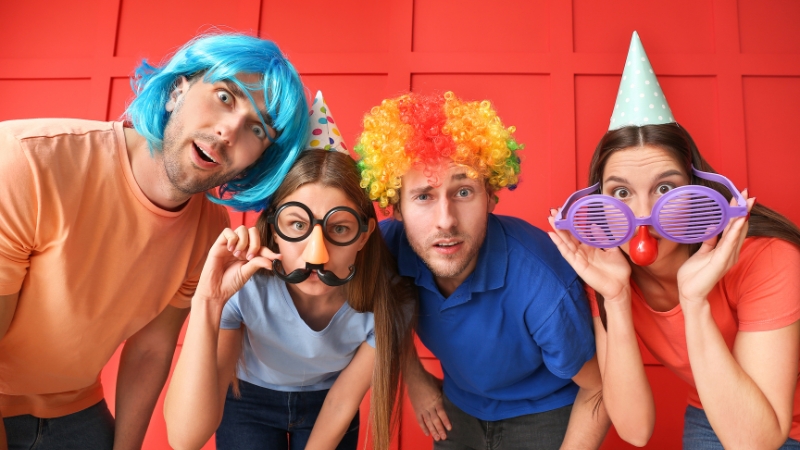 Four people wearing silly costumes, wigs, and party hats posing with exaggerated expressions in front of a red wall
