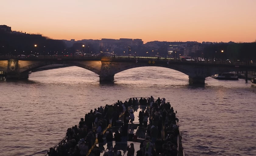 A boat glides down the river in Paris at sunset, with vibrant orange and purple hues reflecting on the water's surface