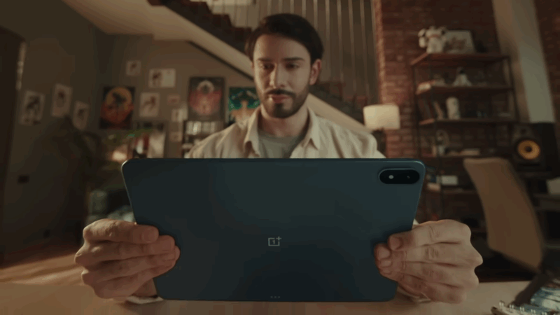 A man holds the OnePlus Pad 3 in landscape mode, showing its slim metal back and rear camera while seated in a modern living room