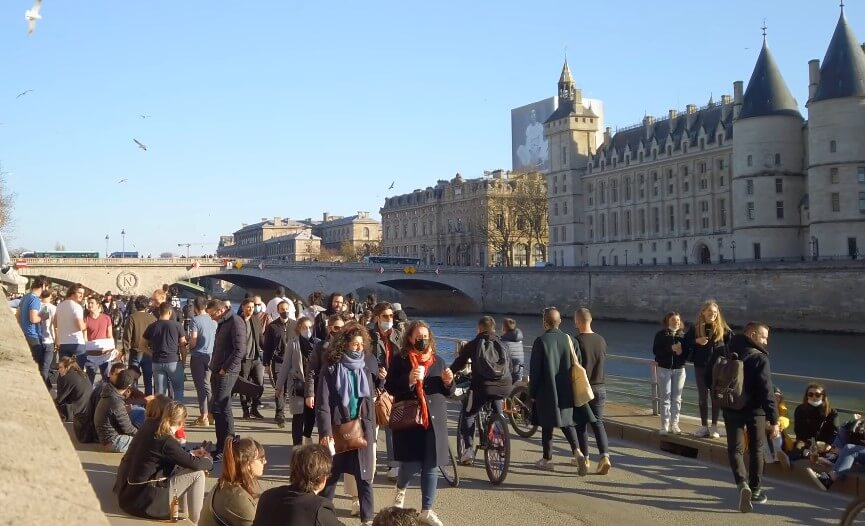 A large crowd of people walking along a river, enjoying the scenery and socializing on a sunny day