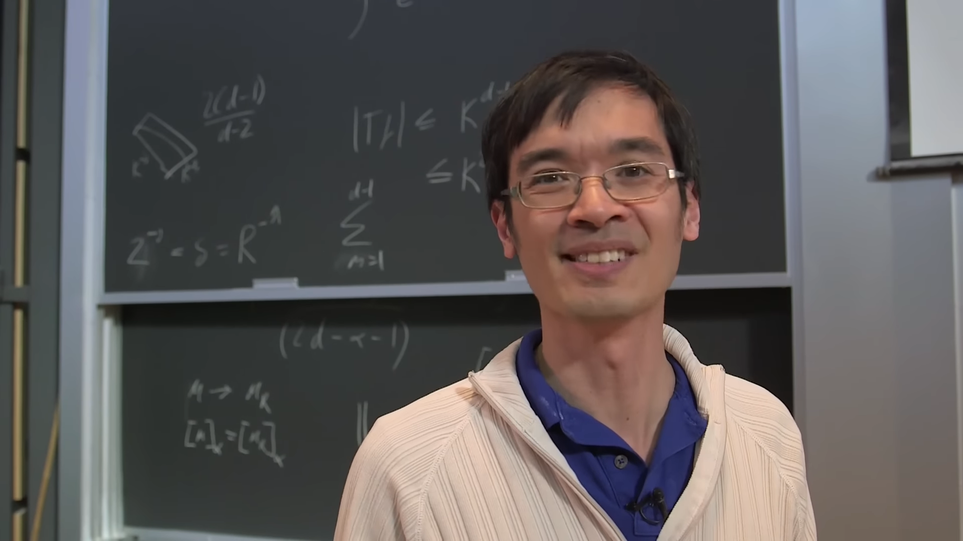 Terence Tao stands in front of a chalkboard filled with equations, smiling during a lecture