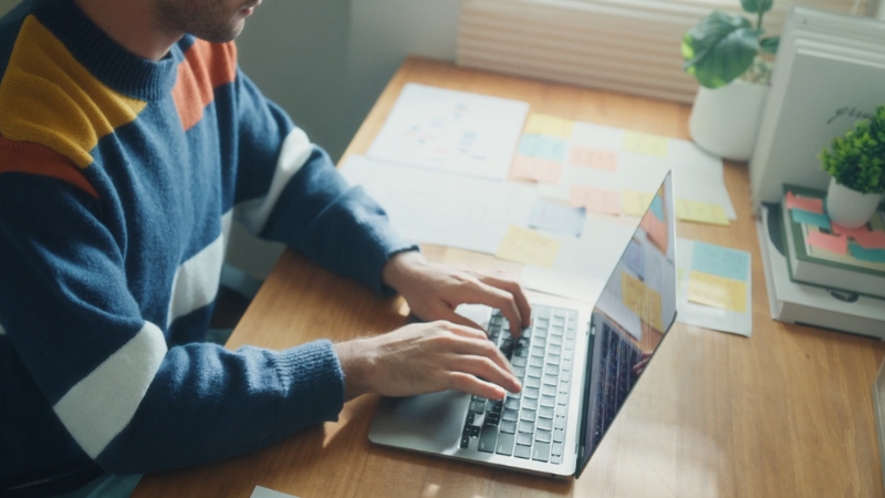 A man types on a laptop at a desk with notes and papers