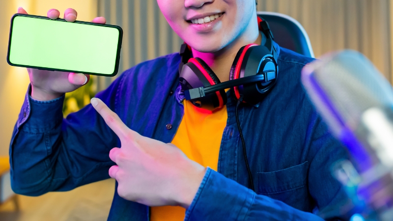 A smiling streamer wearing headphones points to a smartphone with a blank green screen