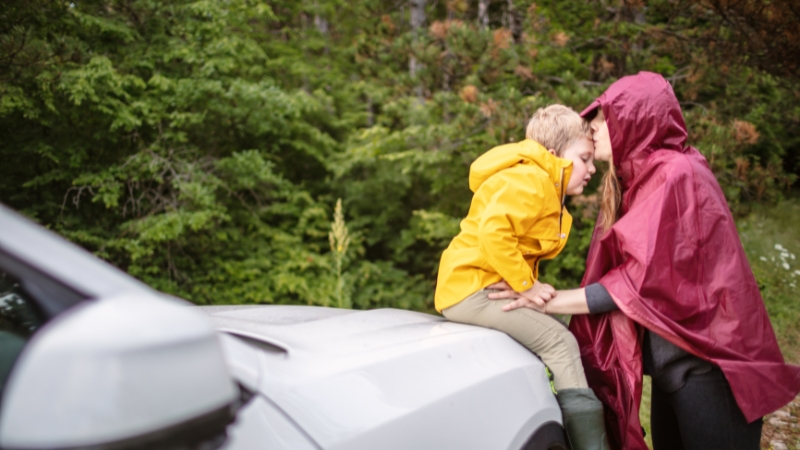 A mum in a rain poncho comforting her child beside a car in nature, capturing the spirit of reliable mum cars for road trips