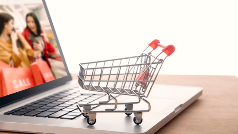 A small shopping cart placed on a laptop keyboard with a sale image on the screen in the background