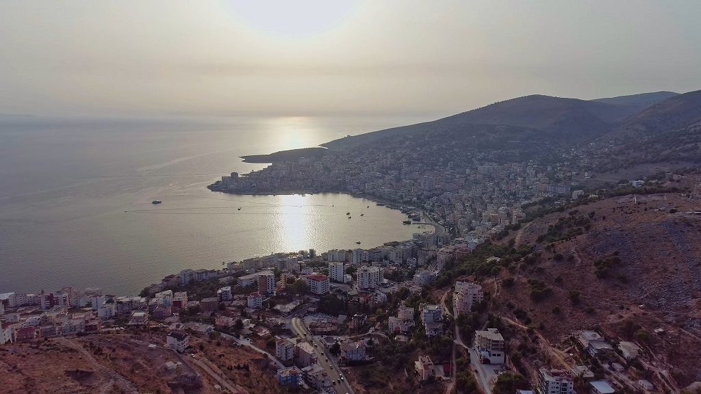 Aerial view of a coastal city in Albania at sunset, with mountains in the background and the Adriatic Sea reflecting golden light
