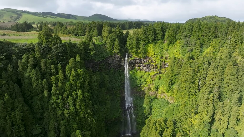 Scenic view of a tall waterfall surrounded by lush green forests and rolling hills in the Azores, Portugal