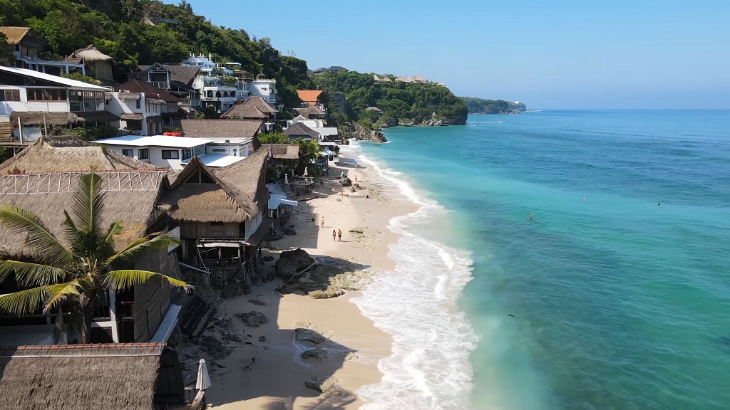 Aerial view of Bali’s coastline with turquoise water, sandy beach, and traditional thatched-roof houses along the shore