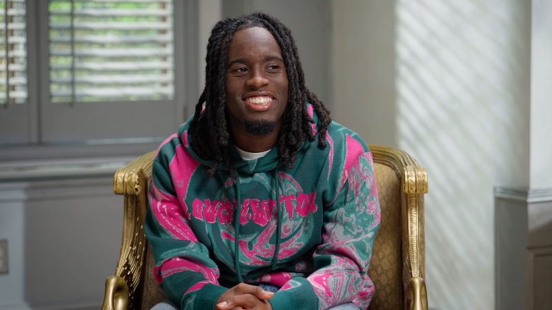 Kai Cenat sitting in a chair wearing a colorful hoodie, smiling during an interview