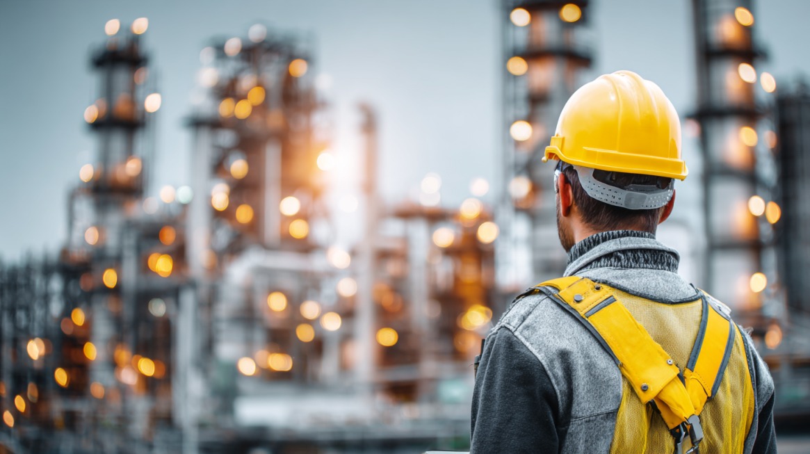 A worker in a yellow hard hat and safety vest standing with their back to the camera, overlooking a blurred oil refinery with glowing lights