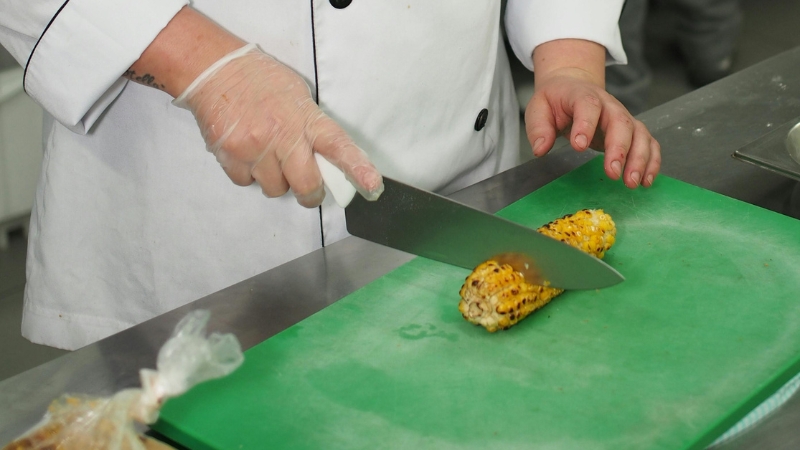 A chef slices grilled corn on a green cutting board