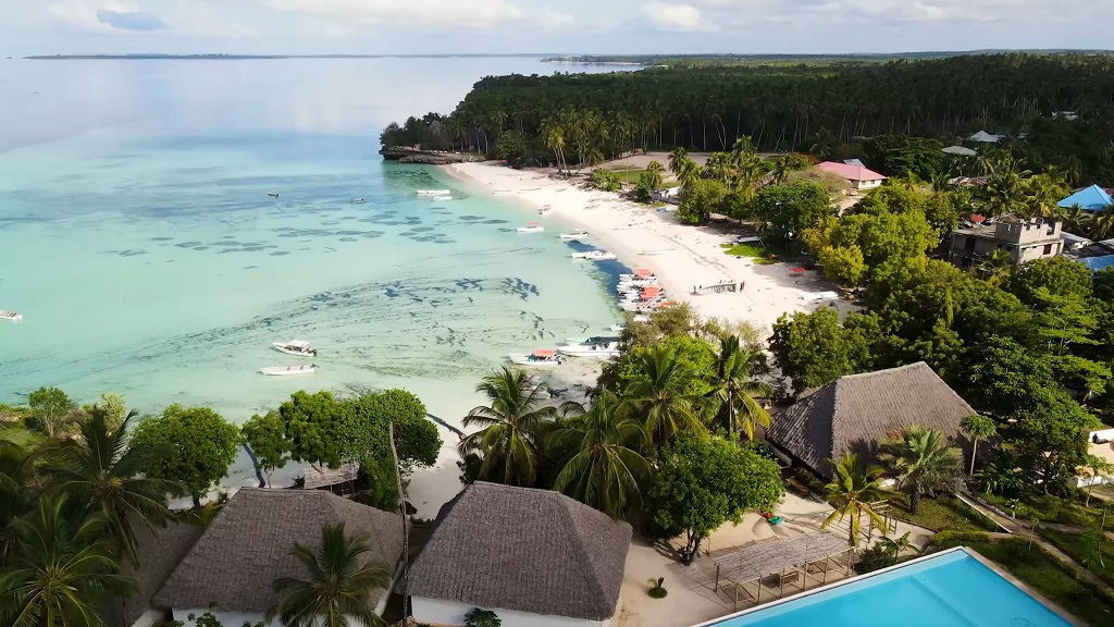 Aerial view of Zanzibar’s coastline with turquoise waters, white sandy beach, palm trees, and thatched-roof bungalows surrounded by tropical greenery