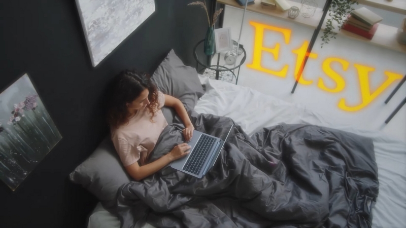 A woman works on a laptop in bed with the word Etsy displayed beside her