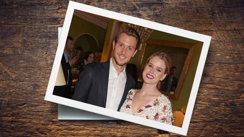 Alex Cowper-Smith and Alice Eve smile together at an indoor event with people in the background