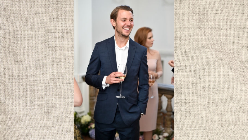 Alex Cowper-Smith in a navy suit smiles and holds a glass at an indoor event