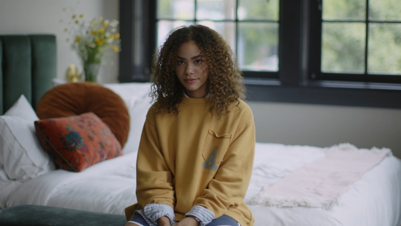 Antonia Gentry sitting on a bed in a cozy room, wearing a yellow sweatshirt