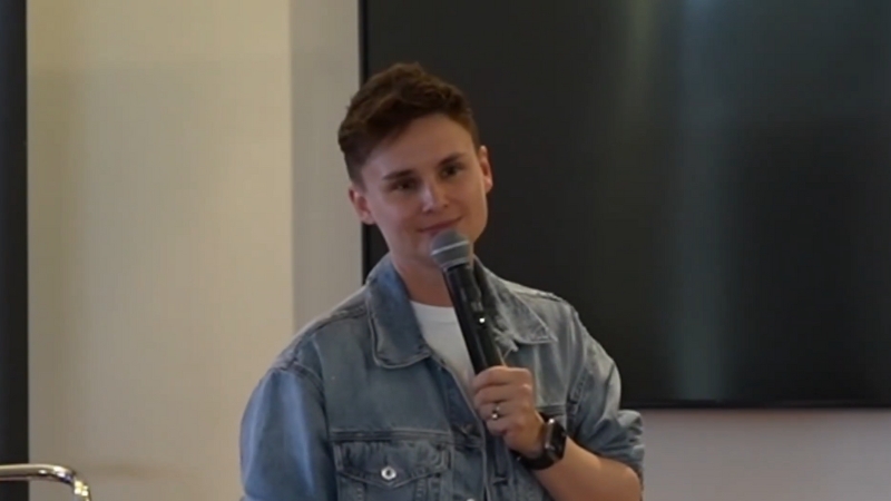 Ashley Gutermuth stands on stage wearing a denim jacket and holding a microphone