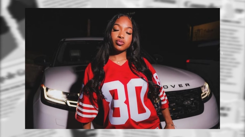 A young woman wearing a red sports jersey stands confidently in front of a white Range Rover at night