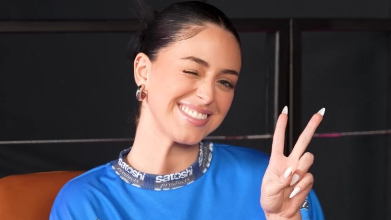 Camilla Araujo smiles and flashes a peace sign in a blue top during an interview