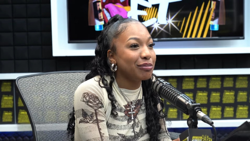 A young woman speaks into a studio microphone during an interview, smiling and wearing layered necklaces and hoop earrings