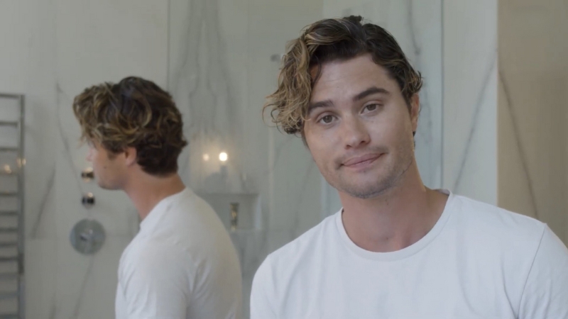 Chase Stokes wears a white t-shirt and looks at the camera with a calm expression in a bright bathroom setting