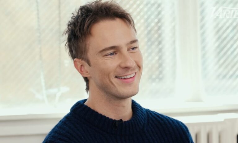 Drew Starkey smiles at the camera while wearing a blue sweater