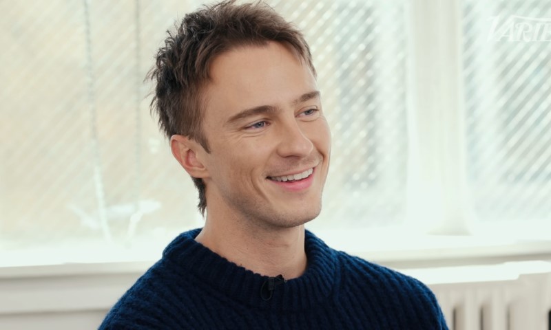 Drew Starkey smiles at the camera while wearing a blue sweater