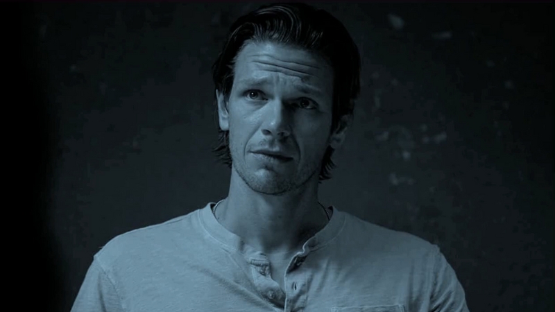 Jon Beavers looks ahead with a serious expression in a dimly lit scene, wearing a casual light shirt