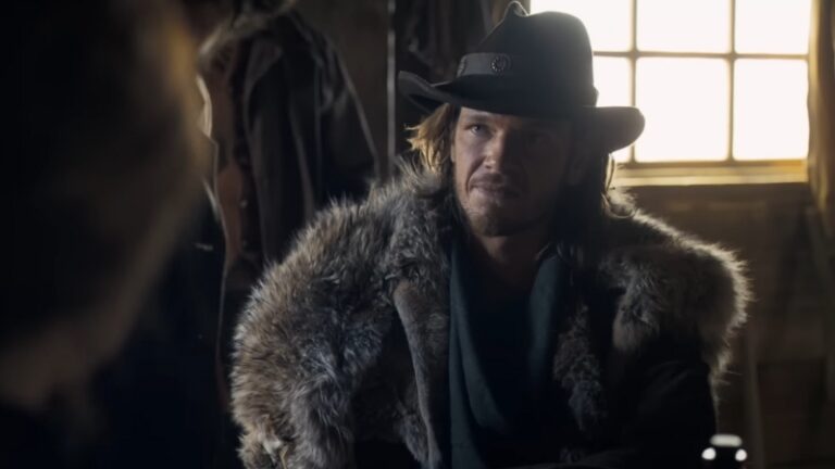 Jon Beavers sits in a dim cabin wearing a fur coat and cowboy hat