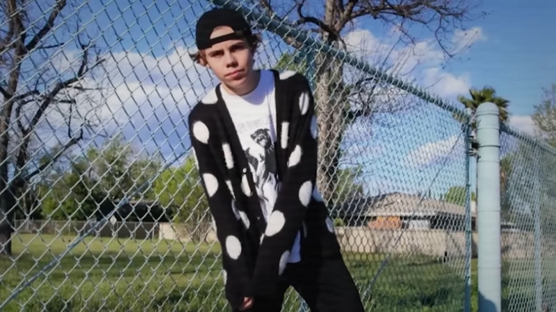 Kid Laroi leans against a chain-link fence wearing a black cap and polka-dot sweater