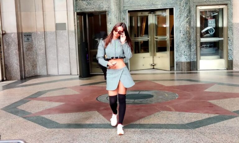 Emily Ratajkowski in a skirt and jacket walks confidently down a well-lit hallway