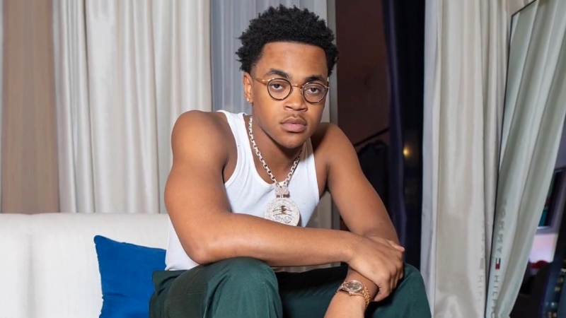 Michael Rainey Jr. with short curly hair, glasses, a white tank top, and a large chain sits on a couch