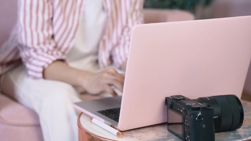 A person works on a pink laptop with a camera placed beside it on the table