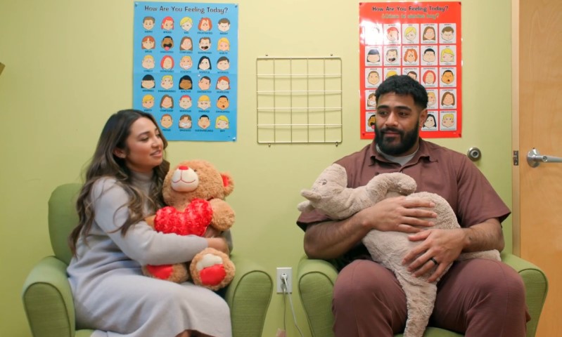 A man and woman sit in chairs, each holding a teddy bear, smiling at each other in a cozy setting