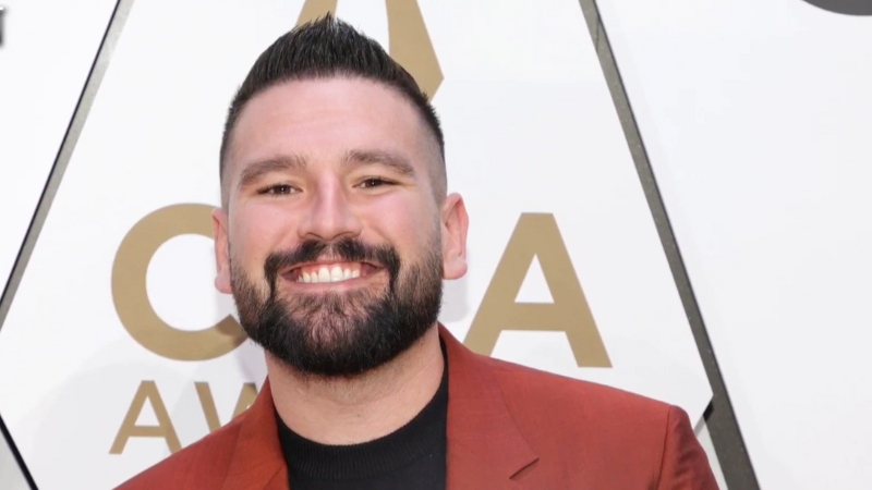 Shay Mooney smiles on the red carpet at an awards event, wearing a red jacket and black shirt