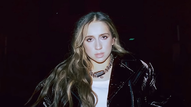 Tate McRae stands at night wearing a shiny black jacket and a chunky necklace