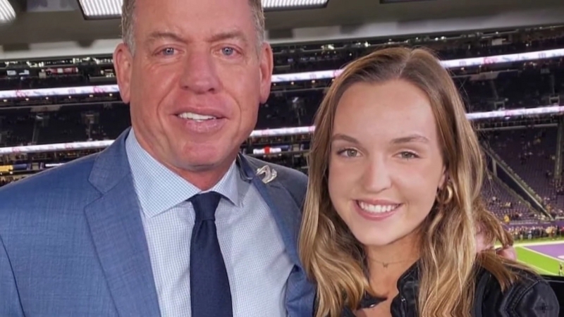 Troy Aikman and Alexa Marie Aikman smile together at a football stadium during a game