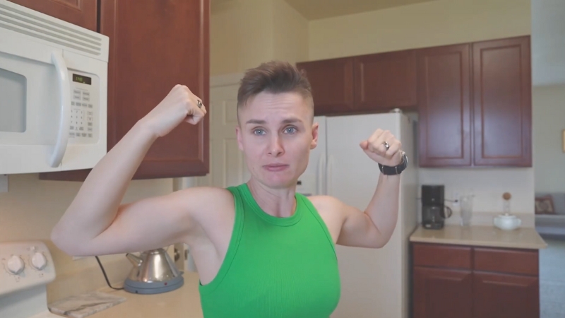 Ashley Gutermuth flexes her arms in a green sleeveless top while standing in a kitchen