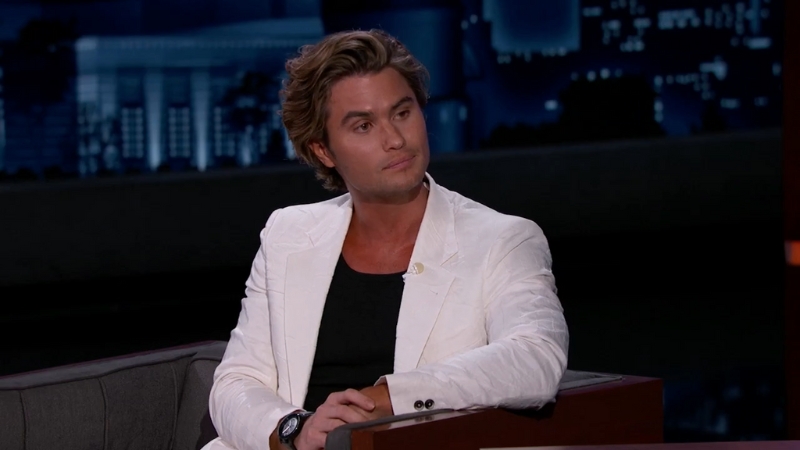 Chase Stokes in a white blazer and black shirt sits on a talk show set