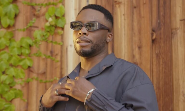 Woody McClain wearing sunglasses and a casual shirt, smiling against a neutral background