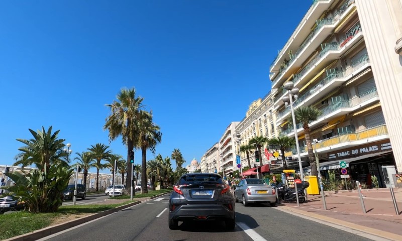A car drives down a street in Nice, lined with tall palm trees under a clear blue sky
