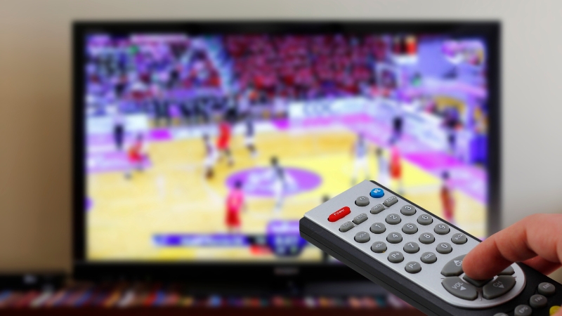 Remote control pointed at a TV showing a live basketball game for Fubo vs DirecTV stream comparison