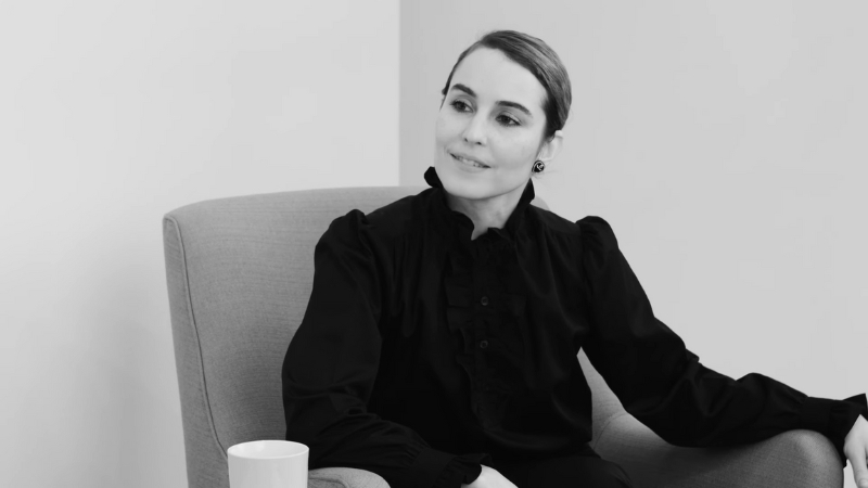 Noomi Rapace sits in a chair in a black outfit during an interview