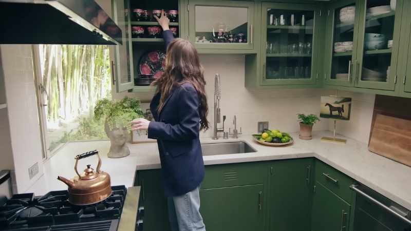 Dakota Johnson reaches for a cup inside her green midcentury kitchen with bamboo visible through the window